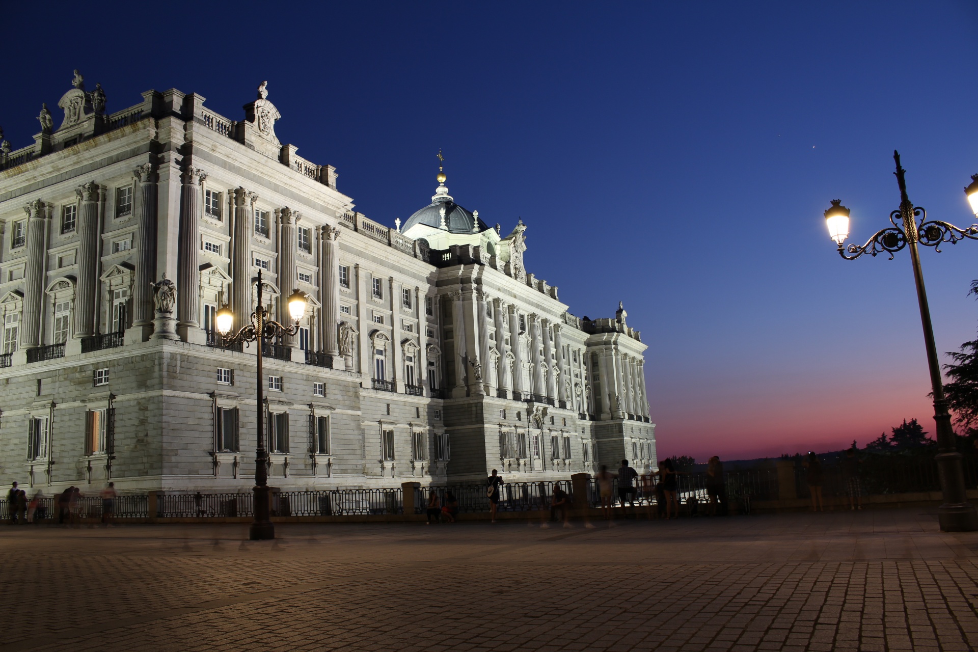 The illuminated Royal Palace of Madrid at night, with its grand stone façade and ornate architectural details glowing against the evening sky.